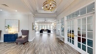 Located at 201 Monticello Mews in Hampton, Virginia, this single-story clubhouse is a thoughtfully designed community gathering space that balances sophistication with comfort. The interior is unified by light-toned wood-look flooring that flows throughout, complemented by crisp white crown molding and neutral wall tones that create a bright, welcoming atmosphere.

The grand entrance foyer draws the eye immediately with a striking gold orb chandelier suspended beneath a coffered ceiling, setting an elegant tone. French-style glass-paned doors line the entry corridor, allowing natural light to filter through while providing visual access to adjoining spaces, including private offices or meeting rooms. A fireplace anchors one lounge area alongside a wall-mounted display screen.

The main lounge opens generously toward a wall of floor-to-ceiling windows and glass doors that flood the space with natural light and offer outdoor views. A vaulted ceiling with a ceiling fan adds architectural interest, while gold pendant lanterns illuminate a bar-height counter adjacent to a well-equipped kitchen featuring quartz countertops, gray cabinetry, stainless steel appliances, and tasteful tile accents.

A dedicated game and recreation wing offers teal-carpeted lounge areas furnished with eclectic seating, a shuffleboard table, foosball table, wall-mounted television, and a casual dining nook—ideal for social gatherings and relaxed entertainment.