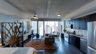 This single-floor urban residence showcases a sleek industrial-chic design that blends raw architectural elements with modern finishes. The exposed concrete ceiling immediately draws the eye upward, lending the space a loft-like character and an open, airy feel. Dark hardwood flooring runs throughout, providing a rich contrast to the lighter tones of the walls and ceiling.

The open-plan layout seamlessly integrates the living, dining, and kitchen areas into one cohesive environment. The living area is anchored by a comfortable sectional sofa, a bold geometric area rug in warm orange tones, and a ceiling fan that adds both function and style. A dining counter with bar seating connects the living space to the kitchen, encouraging a social atmosphere.

The kitchen is well-appointed with navy cabinetry, stainless steel appliances — including a range and over-the-range microwave — and light stone countertops that provide ample workspace.

The standout feature of this residence is undoubtedly the expansive floor-to-ceiling windows that flood the interior with natural light and frame dramatic, unobstructed views of the surrounding city skyline. The result is a sophisticated, contemporary urban living experience that balances industrial character with refined comfort.