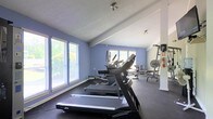 This fitness center offers a practical and functional workout environment designed to support a variety of exercise routines. The space is finished with cool periwinkle-blue accent walls and a crisp white ceiling, giving it a clean and energizing atmosphere. Dark rubber flooring lines the room, providing a durable, slip-resistant surface ideal for high-traffic fitness use.

Multiple treadmills are arranged along the windowed wall, allowing users to enjoy views of surrounding greenery while exercising. Large windows fitted with horizontal blinds line two walls, flooding the space with natural light and creating an open, airy feel. Additional cardio and strength training equipment, including a weight bench and cable machine, are positioned toward the back of the room, offering a well-rounded selection of workout options.

Ceiling-mounted track lighting ensures consistent illumination throughout the space, while a standing fan provides supplemental airflow for comfort during intense workouts. A fire extinguisher is visibly mounted near the entrance, reflecting attention to safety standards. Overall, the fitness center presents a welcoming and well-organized setting suitable for daily exercise routines.