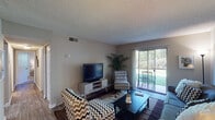 This single-story residence offers a well-organized layout that balances comfort and functionality across its key living spaces. The living room serves as the central gathering area, finished with wood-look flooring, a textured ceiling, and a sliding glass door that opens directly to a private outdoor patio, allowing natural light and views of surrounding greenery to flow inward. Seating areas are arranged around a central coffee table, with space for a media console and television along the wall.

Adjacent to the living room, a dedicated dining area features wood-look flooring, a ceiling fan with light fixture, and a round porthole-style window that adds a distinctive architectural detail. The kitchen is equipped with dark cabinetry, dark countertops, a stainless steel sink, electric range, and a dishwasher, providing a practical and efficient cooking environment.

The unit includes multiple bedrooms, each carpeted and naturally lit through windows with blinds and curtain panels. The primary bedroom benefits from access to both a walk-in closet and a private bathroom. An additional full bathroom features a vanity with a large mirror, a tub-shower combination, and tiled surround. Throughout the unit, neutral-toned walls and textured ceilings contribute to a cohesive and understated aesthetic.
