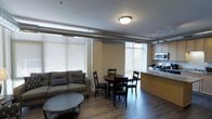 This single-floor space showcases an open-concept layout that seamlessly integrates the living, dining, and kitchen areas into one cohesive environment. The living area is anchored by a comfortable sofa paired with a round coffee table, positioned to take advantage of the natural light streaming through large, horizontal-blind-covered windows along the walls. The dining area features a circular dark wood table surrounded by matching chairs, creating an intimate setting that bridges the living and kitchen zones. The kitchen is defined by a peninsula with a light granite-toned countertop, providing both workspace and a natural partition. It is equipped with stainless steel appliances, including a microwave and stove, with cabinetry in a warm neutral finish above. An exposed cylindrical duct runs along the upper perimeter of the space, lending an industrial character to the otherwise warm interior. Track lighting along the ceiling complements the central flush-mount fixture, ensuring the space is well-lit throughout. The hardwood-style flooring extends seamlessly across the entire area, contributing to a unified and clean aesthetic overall.