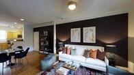 This single-floor space makes a strong design statement through its thoughtful use of contrast and curated décor. The living area serves as the focal point, anchored by a dramatic dark feature wall that sets a rich, moody tone against crisp white trim and ceiling. Warm hardwood flooring runs throughout, lending natural warmth that balances the deeper color palette.

A white sofa takes center stage, adorned with an assortment of textured throw pillows in muted earth tones and warm coral, contributing to an eclectic yet cohesive aesthetic. A patterned area rug in jewel tones grounds the seating arrangement, while a glass-top coffee table keeps the space feeling open and airy. A casual upholstered ottoman adds flexible seating and a relaxed, lived-in feel.

Two large framed abstract artworks hang above the sofa, bringing color and visual interest to the dark wall. A sleek open metal shelving unit near the entryway provides both storage and display space, adding an industrial edge to the overall design. Overhead, a classic flush-mount ceiling light provides ambient illumination.

The overall ambiance is bold yet inviting — a space that balances dramatic design choices with comfortable, functional living in a compact, well-appointed single-level layout.