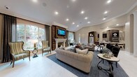 This elegantly designed single-floor space offers an impressive open-plan layout that seamlessly connects multiple living and functional areas. The centerpiece of the main living area is a striking fireplace framed by a reclaimed wood accent wall, topped with a wall-mounted television and flanked by arched windows dressed in floor-length drapes that invite generous natural light. Vaulted and tray ceilings with crown molding add architectural depth, while recessed lighting and a chandelier visible in an adjoining area provide warm, layered illumination throughout. Light-toned large-format tile flooring extends across the space, complementing the neutral warm color palette of the walls and furnishings. Tufted sofas, wingback accent chairs, and decorative coffee tables are thoughtfully arranged to create distinct yet connected seating areas. Structural columns and graceful archways define transitions between zones without interrupting the open flow. Toward the rear, a kitchenette or refreshment area is visible, complete with cabinetry and a stainless steel refrigerator, suggesting the space is well-equipped for hosting. Curated artwork and decorative accents throughout contribute to a polished, welcoming ambiance, making this space ideal for both relaxed gatherings and more formal occasions.