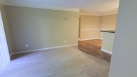 This single-floor space offers a clean, well-maintained interior designed with comfort and functionality in mind. The living area is finished with plush, neutral-toned carpeting that adds warmth underfoot, while a curved transition leads into an adjacent area featuring rich hardwood-style flooring, creating a natural visual distinction between living zones. Soft sage-green and warm beige walls are complemented by crisp white crown molding and baseboards throughout, lending the space a polished and cohesive aesthetic. Recessed lighting is integrated into the ceiling, providing even, ambient illumination across the open layout. A half-wall partition with a dark countertop surface subtly defines the boundary between the main living space and the adjoining area, adding architectural interest without compromising the open feel. Wall-mounted accent lighting adds a subtle decorative touch, while a hallway visible in the background suggests additional rooms beyond the main living area. The overall design is characterized by its neutral color palette, thoughtful detailing, and easy flow between spaces, making it a versatile and inviting environment suitable for a variety of uses.