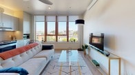 This contemporary single-floor residence offers a thoughtfully designed layout that maximizes both comfort and functionality. The living area is the standout centerpiece of the space, anchored by a large bank of wood-framed windows that flood the room with natural light and frame sweeping views of the surrounding cityscape. Light hardwood flooring runs throughout, complementing the clean, neutral palette of the interior. The living space comfortably accommodates a lounge seating arrangement and features a ceiling fan, creating an airy and relaxed atmosphere.

The kitchen is efficiently designed with light grey cabinetry, a tiled backsplash, stainless steel appliances including a refrigerator and range, and ample counter space for meal preparation. A stainless steel sink and under-cabinet storage complete the practical and modern kitchen layout.

The sleeping area is tucked behind a partition, providing privacy from the main living space, and includes room for a full-sized bed and a desk, making it suitable for both rest and work.

The bathroom features dark slate-style tile flooring, a single vanity with light wood cabinetry, a shower with a curtain enclosure, and a conveniently integrated stacked washer and dryer unit — a practical amenity that enhances the self-sufficiency of the space. Overall, the residence presents a clean, modern aesthetic with urban views and well-appointed finishes throughout.