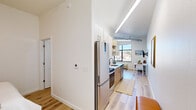 Located at 2600 College Ave in Conway, Arkansas, this efficiently designed one-bedroom, one-bathroom unit offers a thoughtfully planned layout that maximizes every square foot. The open-concept living and kitchen area is anchored by light hardwood flooring that runs throughout, creating a warm and cohesive aesthetic. The kitchen is a standout feature, outfitted with stainless steel appliances including a French door refrigerator, electric range, and over-the-range microwave. Light wood cabinetry paired with white quartz countertops and open shelving gives the kitchen a clean, contemporary feel, while a window at the end of the galley-style run draws in natural light and views of the surrounding neighborhood. Linear LED lighting along the ceiling further brightens the space. The living area flows naturally from the kitchen, offering a comfortable and functional space. The bedroom, though cozy in scale, is a serene retreat with soft neutral tones and warm finishes. The bathroom features a shower-tub combination, a vanity with a stone countertop, and rustic wood floating shelves that add a touch of character. Throughout the unit, crisp white walls, elevated ceilings, and consistent flooring create a bright, modern ambiance well-suited for efficient urban living.