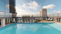 This rooftop amenity level offers a well-appointed collection of social and recreational spaces designed for both relaxation and entertainment. The centerpiece is a generously sized outdoor swimming pool with crystal-clear blue water, surrounded by a sun deck furnished with lounge chairs and a raised wooden deck area, all set against a dramatic urban skyline backdrop that includes prominent downtown high-rises.

A seamless indoor-outdoor transition is achieved through floor-to-ceiling glass walls and large double-pane glass doors that open directly onto the pool terrace. The interior lounge area features light herringbone-patterned hardwood flooring, a decoratively paneled ceiling with geometric detailing, and tall bistro tables with bar seating positioned alongside the glazed facade to maintain views of the outdoor space.

Adjacent to the lounge, a dedicated game room provides entertainment with skee-ball machines, a shuffleboard table, and multiple wall-mounted flat-screen televisions. Slatted wood ceiling accents and warm-toned wall panels give this space a polished yet casual feel.

A connecting corridor with wood-paneled walls and warm ambient lighting links the various zones, with a workstation area also visible. The overall design emphasizes modern aesthetics, abundant natural light, and cohesive indoor-outdoor living enhanced by panoramic city views.