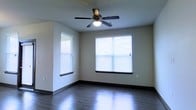 This single-floor space presents a clean, straightforward layout designed for comfort and practicality. The main living area features rich, dark hardwood-style flooring that adds warmth and depth to the room, complemented by smooth neutral-toned walls and dark baseboards that frame the space with subtle contrast. Natural light flows generously through two windows dressed with white horizontal blinds, brightening the interior and creating an airy, open feel. A classic ceiling fan with an integrated light fixture is centrally mounted overhead, providing both ventilation and ambient lighting. The walls are finished in a soft, neutral tone that lends versatility to the space, making it easy to personalize with a variety of décor styles. The overall ambiance is calm and understated, with a focus on clean lines and functional design. The combination of natural light, durable flooring, and simple finishes gives the space a move-in-ready appeal suited for a range of uses.