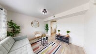 This well-appointed two-floor home showcases a cohesive modern aesthetic, highlighted by light wood-style flooring, crisp white walls, and thoughtfully curated interiors throughout. The welcoming living room offers generous natural light through large windows, complemented by a contemporary multi-arm ceiling light fixture, a comfortable sectional sofa, and a rattan accent chair atop a vibrant striped area rug — creating a warm yet stylish atmosphere. A dedicated dining nook features rattan chairs around a round white table, bathed in natural light from a window overlooking a green outdoor view. The kitchen is a standout space, outfitted with bright white shaker-style cabinetry, granite countertops, a white subway tile backsplash, stainless steel appliances including a dishwasher, and matte black hardware accents throughout. The bedrooms are serene and airy, each featuring light wood flooring, white linens, and tasteful wall art. One bedroom is well-suited for children, accommodating a bunk bed and a small study desk. The bathroom is finished with marble-look tile flooring and wall surround, a granite-topped white vanity, a bold round mirror, and matte black fixtures, adding a refined touch. Overall, the space strikes an inviting balance between modern comfort and clean, functional design.