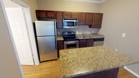 This single-floor residential unit offers a thoughtfully designed layout that balances functionality and comfort. The kitchen serves as a central highlight, outfitted with rich dark-wood cabinetry extending to the ceiling, stainless steel appliances including a refrigerator, electric range, built-in microwave, and dishwasher. Granite countertops with warm gold and brown tones provide generous prep space and carry over to a breakfast bar that opens the kitchen toward the living area, promoting an open, connected feel. Crown molding throughout adds a refined architectural detail, lending the space a polished character. The living area features durable wood-look flooring, neutral warm-toned walls, and a sliding glass door fitted with vertical blinds that allows natural light to filter in and provides access to an outdoor area. A front entry door with a deadbolt opens directly into this welcoming living space. The bathroom is clean and practical, offering wood-look flooring, a granite-topped vanity, a full bathtub with tile surround, a wall-mounted medicine cabinet, and a closet for additional storage. Overall, the space presents a cohesive, move-in-ready environment with consistent finishes and a warm, neutral palette throughout.