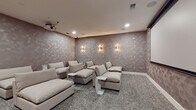 Located at 1420 Lakefront Dr in Sarasota, Florida, this single-floor entertainment space is thoughtfully designed for an immersive cinematic experience. The home theater room is the centerpiece, finished with textured geometric wallcovering in warm taupe tones that enhances the room's acoustic and visual atmosphere. A large retractable projection screen anchors the viewing wall, complemented by a ceiling-mounted projector and built-in surround sound speakers. Plush, oversized modular sofas in neutral tones are arranged for optimal sightlines, accompanied by sleek marble-topped side tables. Elegant gold-toned wall sconces provide soft ambient lighting, while plush speckled carpet underfoot adds comfort and sound absorption. Adjacent to the theater is a well-appointed wet bar corridor, featuring sage green cabinetry with brushed gold hardware, a crisp white quartz countertop, a built-in beverage refrigerator, and a matte black sink. Decorative tile accents the backsplash wall, while a series of woven rattan pendant lights hang overhead, casting a warm, inviting glow. The flooring in this area showcases a distinctive geometric tile pattern in soft blush and white tones. Curated artwork and brass decorative accents complete the refined, boutique aesthetic of this exceptional entertainment retreat.