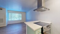 Located at 114 S Gramercy Pl in Los Angeles, CA, this efficient single-floor unit offers a thoughtfully arranged layout comprising a living area, kitchen, bathroom, and closet. The open-concept design seamlessly connects the kitchen and living room, maximizing the sense of space throughout. Light wood laminate flooring runs consistently across the main living areas, lending a warm and cohesive feel to the interior.

The kitchen features a peninsula-style island topped with a white countertop, a built-in electric cooktop, and a stainless steel range hood overhead. The cabinetry is finished in a soft sage green with gold-toned hardware, adding a subtle contemporary accent. A wall-mounted oven completes the cooking area.

The living room is bright and airy, benefiting from natural light that filters in through a large window dressed with horizontal blinds. White walls throughout maintain a clean, neutral backdrop. A wall-mounted HVAC unit and ceiling ventilation ensure climate control within the space.

The bathroom is finished with white square tile surrounding a full soaking tub and shower combination, complemented by matte black fixtures that provide a modern contrast. White marble-style tile floors and a standard vanity with sink complete the room.

A dedicated closet provides convenient built-in storage. Overall, the unit presents a clean, move-in ready aesthetic suited for urban living in the Koreatown neighborhood of Los Angeles.