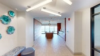 Located along the Lynnway in Lynn, Massachusetts, this single-floor amenity space masterfully blends contemporary design with warm, natural materials to create an inviting and functional gathering environment. The layout flows seamlessly from an open lounge area into a dining and kitchen zone, all unified by consistent light tile flooring and a cohesive design palette.

The lounge area is a standout feature, boasting floor-to-ceiling windows that frame sweeping waterfront views of the harbor. A rich wood-planked ceiling adds warmth overhead, while modern seating arrangements and a wall-mounted television make the space ideal for both relaxation and entertainment. Curated artwork and sculptural accent pieces contribute to the refined, contemporary ambiance.

The dining area centers around a substantial concrete-topped communal table with woven rattan chairs, illuminated by a statement pendant light. Large windows continue to frame the picturesque harbor backdrop, flooding the space with natural light. Adjacent to the dining area, a well-appointed kitchenette features warm wood cabinetry, a dark stone countertop, a sink, and stainless steel appliances, accented by a striking white geometric tile backsplash.

Throughout the space, reclaimed wood accent walls create textural contrast against crisp white surfaces, while a corridor with hexagonal tile detailing and a dual water fountain station adds thoughtful, practical touches to this elegantly designed communal space.