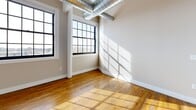 Located at 2075 Kenmore Ave in Buffalo, NY, this 2-bedroom, 2-bathroom loft-style unit showcases an industrial-chic aesthetic with thoughtful modern finishes throughout. The open-concept kitchen and living area anchors the space, highlighted by soaring ceilings with exposed ductwork and ample natural light streaming through large, multi-pane industrial windows. The kitchen is well-appointed with white shaker cabinetry, a granite-topped island with an integrated sink, stainless steel appliances including a refrigerator, range, built-in microwave, and dishwasher, all complemented by stylish pendant lighting. Rich hardwood floors extend throughout the living areas and bedrooms, adding warmth to the open layout. The two bedrooms are generously proportioned, each benefiting from large windows with views of the surrounding neighborhood. Both bathrooms feature clean tile flooring, white vanities with granite countertops, and a full tub-shower combo with built-in shelving. A dedicated laundry room houses a stacked washer and dryer unit for added convenience. A closet provides additional storage. The overall ambiance blends the character of a converted industrial building with the comfort and functionality of a well-designed modern residence.