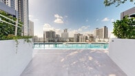 Located in the heart of downtown Miami, this rooftop pool deck offers a refined outdoor retreat with striking panoramic views of the city skyline. The centerpiece is a generously sized lap pool with lane markings, finished in crisp blue tile and surrounded by wide, light-toned pavers that reflect the Florida sunshine. Glass perimeter railings preserve unobstructed sightlines to the surrounding high-rise towers, creating an immersive urban backdrop. A row of contemporary lounge chairs lines the pool's edge, providing ample space for relaxation. Tucked alongside the deck are private cabana structures accented with living green walls, offering shaded retreats framed by tropical plantings including palms and olive trees. Large planter boxes add lush greenery throughout, softening the modern architectural aesthetic. The pool area sits adjacent to the building's glass-and-steel façade with balconied upper floors visible overhead. Whether viewed under bright midday sun or open blue skies, the deck captures the energy of Miami's vibrant urban core while maintaining a calm, resort-like atmosphere. This amenity space is purpose-built for both active use and leisurely enjoyment, with thoughtful landscaping and clean, contemporary design throughout.