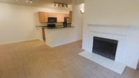 This single-floor space offers an open-concept design that seamlessly connects the living area and kitchen. The living room is anchored by a classic white-mantled fireplace with a tile surround and hearth, adding warmth and a focal point to the space. Soft, neutral-toned carpeting extends throughout the living area, contributing to a comfortable and cohesive aesthetic.

The kitchen is positioned on a slightly elevated platform, separated from the living area by a peninsula counter with dark countertops that provide a striking contrast to the light maple cabinetry above and below. The kitchen is equipped with black appliances, including a built-in microwave and a stainless steel refrigerator, along with a sink set into the peninsula. Track lighting mounted on the ceiling above the kitchen ensures ample task lighting.

The walls throughout are finished in a warm, neutral beige tone, complementing the light cabinetry and carpeting. A wall sconce in the living area adds a subtle decorative touch. The overall ambiance is clean, functional, and move-in ready, with a straightforward layout suited for everyday living.