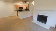 This single-floor space offers an open-concept design that seamlessly connects the living area and kitchen. The living room is anchored by a classic white-mantled fireplace with a tile surround and hearth, adding warmth and a focal point to the space. Soft, neutral-toned carpeting extends throughout the living area, contributing to a comfortable and cohesive aesthetic.

The kitchen is positioned on a slightly elevated platform, separated from the living area by a peninsula counter with dark countertops that provide a striking contrast to the light maple cabinetry above and below. The kitchen is equipped with black appliances, including a built-in microwave and a stainless steel refrigerator, along with a sink set into the peninsula. Track lighting mounted on the ceiling above the kitchen ensures ample task lighting.

The walls throughout are finished in a warm, neutral beige tone, complementing the light cabinetry and carpeting. A wall sconce in the living area adds a subtle decorative touch. The overall ambiance is clean, functional, and move-in ready, with a straightforward layout suited for everyday living.