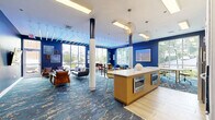 Located at 28 Southfield Ave in Stamford, Connecticut, this thoughtfully designed communal lounge space makes a bold visual statement from the moment you enter. The interior is defined by deep navy blue walls that create a rich, sophisticated atmosphere, complemented by a striking abstract-patterned carpet in oceanic shades of blue and silver that flows throughout the space.

Architectural interest is provided by structural columns and a distinctive ceiling feature — parallel horizontal slats backlit with vivid blue LED lighting — that adds a modern, dynamic quality to the room. Large floor-to-ceiling windows along multiple walls flood the space with natural light while offering open views of the surrounding Stamford streetscape.

The layout is thoughtfully zoned to serve multiple purposes. A comfortable lounge area features a mix of navy blue sofas, rich cognac leather chairs, and coffee tables, creating an inviting setting for relaxed gatherings. Framed nautical-inspired artwork decorates the walls alongside a wall-mounted flat-screen television. A dedicated dining and bar area includes counter seating with sleek stools and small bistro-style tables.

A kitchenette anchors one end of the space, equipped with white cabinetry, a stainless steel refrigerator, a built-in microwave, and a central island with a white countertop. Warm gold geometric pendant lights add an elegant finishing touch to this functional yet stylish amenity space.