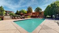 Located at 4601 Montaño Rd NW in Albuquerque, New Mexico, this outdoor community space showcases a thoughtfully designed recreation area that embodies the warmth and character of Southwestern architecture. At the heart of the space is a generously sized swimming pool with clear, blue-tiled water, surrounded by an expansive concrete pool deck that provides ample room for relaxation and recreation.

The pool area is well-appointed with multiple lounge chairs and seating arrangements, complemented by colorful market umbrellas that offer shade throughout the day. A clubhouse building anchors one side of the pool, featuring a terracotta tile roof, stucco exterior, and large glass-panel doors that open directly onto the deck — a classic adobe-inspired design that blends seamlessly with the New Mexico landscape.

The grounds are lush and mature, with towering trees, well-maintained shrubs, and decorative plantings that provide natural shade and greenery throughout the space. Raised planter beds with ornamental grasses and flowering plants add layers of visual interest to the hardscaped areas. Tiered terraces with low stucco walls and wrought-iron fencing create distinct zones within the space, including quieter seating nooks ideal for relaxation. A pergola-covered lounge area adds an additional shaded gathering spot. The entire space is bathed in abundant New Mexico sunshine, creating an inviting, resort-like atmosphere year-round.