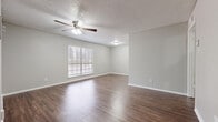 This single-floor unit is finished with dark hardwood-style flooring that runs consistently throughout every room, providing a cohesive and low-maintenance foundation. The living area is generously proportioned, featuring a ceiling fan with an integrated light fixture, a textured popcorn ceiling, and a large window fitted with horizontal blinds that admits natural light. Neutral gray walls and clean white baseboards contribute to a bright, understated aesthetic throughout.

The layout, as seen in the overhead model, includes a living area that flows toward a galley-style kitchen. The kitchen is equipped with white cabinetry, a full-size refrigerator, an electric range, and an over-the-range microwave. A mosaic tile backsplash adds a subtle decorative detail. A door off the kitchen leads to an additional room or utility space.

The bedroom is a well-sized room with its own ceiling fan, a window with blinds, and a door leading to the hallway. The bathroom features a full soaking tub with white shiplap-style surround, a toilet, and towel bars. Ventilation is provided by ceiling-mounted HVAC registers visible throughout the unit.
