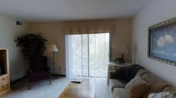 This single-floor space features a living area centered around a large sliding glass door dressed with vertical blinds and a fabric valance, allowing natural light to fill the room while offering views of surrounding greenery. The layout accommodates a mix of seating, including a cushioned sofa and a classic wingback chair, alongside a round side table and floor lamps that contribute to a warm, functional ambiance. The flooring transitions between carpet and hard surface near the sliding door. A textured ceiling adds a traditional character to the space. The sliding glass door provides direct access to an outdoor area, making it a practical and inviting feature of the living space.