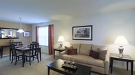 This two-floor residential space offers a comfortable and functional layout designed for everyday living. The living area is finished with soft wall-to-wall carpeting and painted in neutral tones that create a warm, inviting atmosphere throughout. A large window dressed with decorative rust-colored curtains allows natural light to brighten the room, contributing to an open and airy feel.

The living room is thoughtfully arranged with ample space for seating and relaxation. A cushioned sofa with accent pillows serves as the focal point of the room, complemented by a dark wood coffee table and a side table with a classic table lamp that adds a soft, ambient glow. Framed artwork on the wall adds a finishing touch to the clean, understated décor.

Adjacent to the living room, the dining area accommodates a dark wood table and chairs, making it ideal for everyday meals or casual gatherings. The open flow between the living and dining spaces enhances the sense of roominess and connectivity within the home.

Overall, the space presents a tidy, well-maintained interior with a neutral color palette, practical furnishings, and a layout that balances comfort with functionality across two floors.
