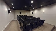 Located at 280 New Haven Blvd in Montgomery, Alabama, this dedicated private home theater is designed to deliver an immersive cinematic experience. The room features a tiered, stadium-style seating layout with two rows of black theater-style seats, each equipped with built-in cup holders and armrests, ensuring comfort for every viewer. An ornate wrought iron railing elegantly separates the raised rear seating platform from the lower front section, adding a refined architectural detail to the space.

The ceiling is finished in a deep matte black, giving the room a true theater atmosphere and minimizing light reflection. Recessed ceiling lights provide controlled ambient illumination, while a ceiling-mounted projector delivers crisp imagery onto a large projection screen positioned along the front wall, which is accented with a dark lower panel for contrast and visual depth.

The surround sound system is prominently featured, with multiple wall-mounted speakers distributed throughout the room for an enveloping audio experience. Soft wall sconces add a warm, cinema-lobby ambiance along the side and rear walls. A built-in projection booth window is visible at the rear, adding an authentic theatrical touch. Neutral carpet flooring and light-toned walls complete this well-appointed, purpose-built screening room.