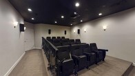 Located at 280 New Haven Blvd in Montgomery, Alabama, this dedicated private home theater is designed to deliver an immersive cinematic experience. The room features a tiered, stadium-style seating layout with two rows of black theater-style seats, each equipped with built-in cup holders and armrests, ensuring comfort for every viewer. An ornate wrought iron railing elegantly separates the raised rear seating platform from the lower front section, adding a refined architectural detail to the space.

The ceiling is finished in a deep matte black, giving the room a true theater atmosphere and minimizing light reflection. Recessed ceiling lights provide controlled ambient illumination, while a ceiling-mounted projector delivers crisp imagery onto a large projection screen positioned along the front wall, which is accented with a dark lower panel for contrast and visual depth.

The surround sound system is prominently featured, with multiple wall-mounted speakers distributed throughout the room for an enveloping audio experience. Soft wall sconces add a warm, cinema-lobby ambiance along the side and rear walls. A built-in projection booth window is visible at the rear, adding an authentic theatrical touch. Neutral carpet flooring and light-toned walls complete this well-appointed, purpose-built screening room.