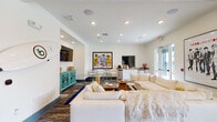 This single-floor communal space is thoughtfully designed to encourage gathering and relaxation, blending contemporary style with welcoming, residential comfort. The layout flows seamlessly between distinct functional zones, each unified by a crisp white color palette punctuated by bold teal accent walls that add energy and visual interest throughout.

The dining area serves as a central focal point, anchored by a large marble-top table surrounded by upholstered gray chairs with nailhead trim. A tray ceiling with a pendant light overhead adds architectural definition to this zone. Adjacent to the dining area, a built-in white cabinetry unit with granite countertops sits beneath a wall-mounted flat-screen television, providing a practical and stylish serving or display surface.

French doors line one wall, flooding the space with natural light and offering views of an outdoor balcony beyond. The lounge area features generously sized white sectional sofas, layered rugs, and a wood coffee table, creating a relaxed setting ideal for socializing. A teal credenza with decorative fretwork supports a second wall-mounted television, while bold pop-art prints add personality throughout. A kitchenette with a dishwasher, coffee station, and countertop space supports the space's entertaining function.