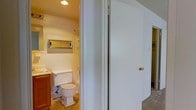 This single-floor space offers a functional and straightforward layout centered around a well-appointed bathroom. The bathroom is finished with classic white subway tile walls that extend partway up, lending a clean and timeless aesthetic to the room. Recessed ceiling lighting provides bright, even illumination throughout the space. A mirrored medicine cabinet is mounted above the vanity area, offering both practical storage and added visual depth. The vanity features warm wood cabinetry paired with a white countertop, creating a subtle contrast against the tiled walls. A standard toilet is positioned centrally within the bathroom, and the flooring features a neutral, textured tile that complements the overall palette. Adjacent to the bathroom is a hallway with a white-paneled door fitted with a brass knob, suggesting additional rooms or areas beyond. The carpeted hallway area adds a layer of warmth and comfort to the transitional space. Overall, the space presents a practical, well-maintained interior with classic finishes and a cohesive, understated design throughout.