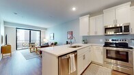 This single-floor residence is thoughtfully designed with a clean, contemporary aesthetic and a functional open-concept layout. The kitchen serves as a central hub, featuring white shaker cabinetry, a gray subway tile backsplash, quartz countertops, and a peninsula island with seating. Stainless steel appliances — including a French door refrigerator, range, microwave, and dishwasher — complete the well-equipped space. Warm hardwood-style flooring flows throughout the main living areas, connecting the kitchen seamlessly to the living room.

The living room is bright and welcoming, benefiting from large sliding glass doors that invite natural light and provide access to an outdoor balcony. Soft, neutral wall tones create a calm atmosphere throughout.

The bedroom offers plush carpet flooring, a ceiling fan, and a large window with views of the surrounding building exterior, filling the room with additional natural light. The bathroom is finished with a white vanity offering ample storage, a wide mirror, and a shower-tub combination with white tile surround.

A dedicated in-unit laundry closet with a stacked washer and dryer adds everyday convenience, rounding out this well-appointed and efficiently designed residence.