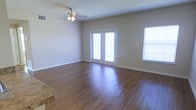 This single-story space presents a welcoming and open living area defined by rich, dark hardwood flooring that runs throughout, adding warmth and a polished aesthetic to the interior. The room is bathed in natural light, courtesy of a set of French doors that lead to the exterior and an additional side window fitted with blinds, ensuring both brightness and privacy control. The neutral, beige-toned walls provide a versatile backdrop that complements a wide range of furnishing styles and décor choices. A ceiling fan with an integrated light fixture is centrally mounted, contributing to both air circulation and ambient lighting. The clean lines of the white door and window trim offer a crisp contrast against the warm wall tones, adding a subtle architectural detail to the space. A glimpse of granite countertop visible at the edge of the frame suggests an adjacent kitchen or bar area, hinting at a practical and connected layout. The open floor plan creates a sense of generous, unobstructed space, making it well-suited for a variety of living arrangements. Overall, the space exudes a simple, tidy, and functional character with thoughtful natural light integration.