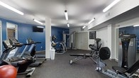 This single-floor fitness center offers a comprehensive workout environment designed to support a wide range of exercise routines. The space is finished with durable rubber flooring, providing a safe and practical surface for both cardio and strength training activities. Bright, elongated fluorescent lighting fixtures are suspended from the white ceiling, ensuring the entire area is well-lit throughout the day. A bold blue accent wall adds a striking visual element to the room, complementing the clean white walls and structural columns throughout the space. The cardio section includes treadmills and an elliptical trainer, while the strength training area features a cable machine, a weight bench, and a seated resistance machine, offering users a full-body workout experience. A wall-mounted flat-screen television is positioned for easy viewing during workouts. A posted fitness club rules sign near the entrance reflects a commitment to maintaining an organized and respectful environment. The layout is thoughtfully arranged to maximize usable floor space while allowing comfortable movement between equipment. Overall, the fitness center presents a clean, functional, and motivating atmosphere suited for a variety of fitness levels and training goals.