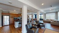 Located along Terrace Greene Circle in Barboursville, Virginia, this single-story space is designed as an open, multi-functional gathering area ideal for communal use. The layout flows seamlessly from zone to zone, unified by warm hardwood flooring and crown molding throughout.

The kitchen is well-appointed with rich honey-toned wood cabinetry, dark granite countertops, stainless steel appliances including a full-size refrigerator, microwave, and range, and generous counter space along a galley-style wall. A central island with bar seating provides an informal gathering point adjacent to the kitchen.

The main lounge area is expansive, accommodating multiple distinct seating arrangements—including leather club chairs around a coffee table and plush sofas on area rugs—allowing for flexible use. Structural columns with decorative detailing define zones without closing off the open floor plan. Natural light enters through multiple windows dressed with white blinds, brightening the neutral gray and taupe walls.

A dedicated dining nook features a round wood table with seating for several guests and a wall-mounted television. A secondary, more intimate sitting room with its own wall-mounted TV and sectional sofa offers a quieter retreat within the space. Recessed lighting and crown molding add a polished, finished quality throughout.
