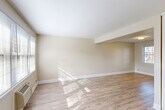 Located at 598 Bullocks Point Ave in East Providence, Rhode Island, this single-floor leasing office serves as the welcoming hub for Bullocks Point Village, an apartment community managed by The Simon Companies. The interior is thoughtfully designed with warm wood-look flooring, neutral gray walls, and recessed lighting throughout, creating a bright and professional atmosphere.

The main reception area doubles as a meeting space, featuring a large wood conference table surrounded by modern chairs, a comfortable seating area, and a reception desk tucked into a windowed alcove. Sheer white curtains filter abundant natural light from large windows, and contemporary abstract artwork adds a decorative touch to the walls.

Two private offices provide dedicated workspace for leasing staff. One office is furnished with a rich wood executive desk, an ergonomic chair, and bold abstract wall art, while a circular mirror and natural greenery add character to the space. A connecting hallway links the offices and includes access to additional utility areas.

The exterior of the property is equally inviting, with vibrant seasonal landscaping, mature trees, and well-maintained grounds surrounding the brick residential buildings of the community. The office entrance features a clear view of the Bullocks Point Village signage, establishing a strong sense of arrival for prospective and current residents.