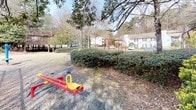 This residential community's outdoor spaces are defined by a well-established natural landscape, featuring mature deciduous trees and dense evergreen shrubs that create a layered, verdant environment throughout the grounds. The buildings present a traditional architectural style with gray siding, covered decks, and exterior staircases, offering residents elevated outdoor areas that overlook the surrounding greenery.

The communal grounds include a dedicated playground area equipped with classic play structures, including a seesaw and swing set frames finished in bold red, set within an open grassy area bordered by neatly trimmed hedgerows. Tall pines and hardwoods frame the space, providing natural shade and a sense of enclosure. A tennis court enclosed by chain-link fencing is also visible on the property, adding to the recreational amenities available to residents.

Landscaping throughout the grounds incorporates raised timber edging, mulched beds, and a variety of shrubs that contribute to the community's maintained yet naturalistic appearance. The open layout between buildings allows for generous spacing and views of the wooded surroundings, giving the community a quiet, park-like atmosphere that blends structured outdoor amenities with natural greenery.
