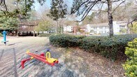 This residential community's outdoor spaces are defined by a well-established natural landscape, featuring mature deciduous trees and dense evergreen shrubs that create a layered, verdant environment throughout the grounds. The buildings present a traditional architectural style with gray siding, covered decks, and exterior staircases, offering residents elevated outdoor areas that overlook the surrounding greenery.

The communal grounds include a dedicated playground area equipped with classic play structures, including a seesaw and swing set frames finished in bold red, set within an open grassy area bordered by neatly trimmed hedgerows. Tall pines and hardwoods frame the space, providing natural shade and a sense of enclosure. A tennis court enclosed by chain-link fencing is also visible on the property, adding to the recreational amenities available to residents.

Landscaping throughout the grounds incorporates raised timber edging, mulched beds, and a variety of shrubs that contribute to the community's maintained yet naturalistic appearance. The open layout between buildings allows for generous spacing and views of the wooded surroundings, giving the community a quiet, park-like atmosphere that blends structured outdoor amenities with natural greenery.