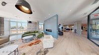 This generously sized amenity space in Durham, NC offers a thoughtfully designed single-floor layout that blends contemporary style with comfortable, multi-functional living areas. Light hardwood flooring runs throughout, anchoring the open, airy atmosphere created by an abundance of natural light pouring in through floor-to-ceiling windows and glass walls that frame sweeping views of the surrounding cityscape.

The first living area serves as an elegant lounge, anchored by a sleek linear fireplace set against a bold blue accent wall. Exposed concrete columns add an industrial touch, while pendant lighting and curated seating arrangements — including sectional sofas and accent chairs atop colorful area rugs — create an inviting social environment. A glass-enclosed nook with outdoor views provides an intimate setting for smaller gatherings.

The second living area shifts to a more recreational tone, featuring a pool table, foosball table, and casual lounge seating alongside a wall-mounted display screen — ideal for entertainment and community activities. A dining counter with bar seating, wood slat ceiling details, and a dark-framed glass stairwell enclosure add architectural interest. A hallway connects the zones seamlessly, completing this well-appointed, versatile community amenity space.