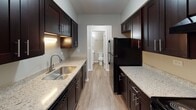 Located at 23435 S Western Ave in Park Forest, Illinois, this single-story unit offers a well-organized layout with a clean, updated aesthetic throughout. The floor plan reveals distinct living, sleeping, and utility zones connected by a central corridor.

The living area is generously proportioned, featuring neutral-toned carpet underfoot and warm beige walls. Natural light enters through a window fitted with blinds, and a baseboard heating unit runs along the wall. The transition from the living space into the entry and hallway areas is marked by wood-look vinyl plank flooring, which continues through the kitchen and bathroom zones, providing a cohesive and durable surface.

The kitchen is a notable highlight, featuring a galley-style design with rich dark cabinetry, granite-look countertops, an undermount stainless steel sink, and black appliances. Ample upper and lower cabinet storage lines both sides of the kitchen, maximizing functionality.

The bedrooms are carpeted and include closets with bifold doors, offering practical storage. Windows bring in natural light and views of the surrounding exterior.

The bathrooms are well-appointed: one features a white vanity with chrome fixtures and a lighted mirror, while the other includes a full tub-and-shower combination with floor-to-ceiling dark tile surround and chrome hardware. An exposed brick accent wall near the entry adds a distinctive architectural character to the space.
