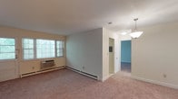 This single-floor residence at Chelsea Village, No. 88, offers a straightforward and functional layout across several well-defined spaces. The living area is generously proportioned, finished with neutral-toned wall-to-wall carpeting and painted in soft, light tones that contribute to a calm and airy atmosphere. A wall-mounted air conditioning unit and baseboard heating ensure year-round comfort, while windows allow natural light to filter in and provide views of surrounding greenery.

The kitchen is a notable feature, equipped with rich cherry-toned cabinetry, granite-style countertops, and dark tile backsplash. Black appliances—including a gas range, microwave, and dishwasher—complement the warm cabinetry tones, and a window above the sink brings in additional natural light. Light tile flooring completes the practical yet attractive space.

The bedroom areas are carpeted and neutral, offering clean, versatile spaces with closet access. A dedicated entry area includes an additional closet for storage. The bathroom features a full tub and shower combination enclosed by sliding glass doors, set against beige tile surrounds with a clean, understated finish. Overall, the residence presents a cohesive, move-in-ready environment.