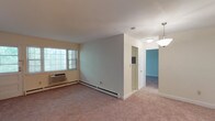This single-floor residence at Chelsea Village, No. 88, offers a straightforward and functional layout across several well-defined spaces. The living area is generously proportioned, finished with neutral-toned wall-to-wall carpeting and painted in soft, light tones that contribute to a calm and airy atmosphere. A wall-mounted air conditioning unit and baseboard heating ensure year-round comfort, while windows allow natural light to filter in and provide views of surrounding greenery.

The kitchen is a notable feature, equipped with rich cherry-toned cabinetry, granite-style countertops, and dark tile backsplash. Black appliances—including a gas range, microwave, and dishwasher—complement the warm cabinetry tones, and a window above the sink brings in additional natural light. Light tile flooring completes the practical yet attractive space.

The bedroom areas are carpeted and neutral, offering clean, versatile spaces with closet access. A dedicated entry area includes an additional closet for storage. The bathroom features a full tub and shower combination enclosed by sliding glass doors, set against beige tile surrounds with a clean, understated finish. Overall, the residence presents a cohesive, move-in-ready environment.