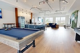 This 1,130 square foot single-floor space in Katy, TX, is a meticulously designed communal area that seamlessly blends functionality with style. The room boasts an open floor plan with high ceilings and large windows, allowing natural light to flood the space. The polished wood flooring adds warmth and elegance throughout.

The area is equipped with multiple entertainment options, including two pool tables and a shuffleboard table, perfect for casual gaming and socializing. A comfortable lounge area features a plush blue sectional sofa and modern accent chairs, ideal for relaxation or casual conversations.

The space also includes a fully-equipped bar with sleek blue cabinetry, a white countertop, and bar stools, creating an inviting spot for refreshments. A commercial-grade refrigerator behind the bar ensures drinks stay cool.

Artistic elements add character to the room, with framed vintage-style beverage posters and a striking living wall featuring cascading artificial flowers and a neon "Live it Love it" sign. Track lighting and unique pendant fixtures provide ambient illumination.

Large glass doors and windows offer views of the surrounding area, connecting the interior to the outdoor environment. This versatile space is designed to accommodate various social activities, making it an ideal gathering spot for residents or community members.