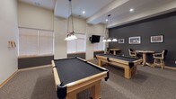 This single-floor recreation and lounge space is thoughtfully designed for social gatherings and leisure activities. The centerpiece of the room is two full-size pool tables with black felt surfaces, each illuminated by decorative pendant lights suspended from the elevated, vaulted ceiling. The generous ceiling height adds an open, airy quality to the entire space, enhancing its welcoming atmosphere.

A wall-mounted flat-screen television is positioned for easy viewing throughout the room, while a bistro-style high-top table with bar chairs offers a casual spot for conversation or games. A dedicated lounge area features multiple dark upholstered armchairs arranged around a potted palm plant, creating a comfortable retreat within the larger space. Framed artwork adorns the walls throughout, adding a refined touch to the recreational setting.

The room is finished with patterned carpet flooring and features a two-tone wall treatment, with a darker charcoal accent on the lower portion and neutral tones above. Large windows fitted with horizontal blinds allow natural light to filter into the pool table area. Recessed lighting throughout ensures the space remains well-lit. A secondary access door connects to an adjoining area, contributing to the overall functionality and flow of this well-appointed recreational facility.