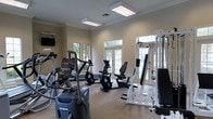 This single-story fitness center offers a thoughtfully designed workout environment that balances functionality with a polished aesthetic. The room features elegant crown molding along the ceiling, adding a refined architectural touch rarely seen in gym spaces. Overhead, recessed fluorescent light panels provide bright, even illumination throughout the area.

Natural light plays a prominent role in the ambiance, streaming in through multiple large windows and a set of classic French doors that face the exterior, offering views of the surrounding greenery. This combination of artificial and natural lighting creates an open, energizing atmosphere well-suited for exercise.

The space is outfitted with a comprehensive selection of fitness equipment, including an elliptical trainer, a recumbent stationary bike, an upright cycle, and a multi-station cable strength machine, providing options for both cardiovascular and resistance training. A wall-mounted television is positioned for easy viewing during workouts.

The neutral-toned walls and light-colored flooring contribute to a clean, airy feel, while the organized layout allows for comfortable movement between machines. Overall, the fitness center presents a welcoming and practical environment for a well-rounded workout routine.
