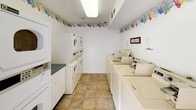 This shared laundry facility is a single-story space designed for practical, everyday use. The interior is bright and clean, with white walls accented by a cheerful laundry-themed wallpaper border that runs along the perimeter near the ceiling, adding a touch of character to the functional environment. The flooring throughout consists of durable stone-look ceramic tiles in warm neutral tones.

The main laundry area is well-stocked with multiple top-loading commercial washing machines arranged in a row, complemented by stacked washer-dryer units along the adjacent wall, providing ample capacity for shared use. A dedicated folding counter with white cabinetry below offers a convenient workspace. A paper towel dispenser and posted usage guidelines are thoughtfully included for user convenience.

The facility is illuminated by overhead fluorescent lighting, ensuring a well-lit environment at all times. Rolling wire laundry carts are available for transporting clothes with ease. Access is controlled via a keypad-secured glass-panel door, enhancing security. A second glass-panel door offers an additional entry and exit point, both allowing natural light to filter in. A wet floor sign reflects the facility's commitment to cleanliness and safety.