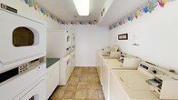 This shared laundry facility is a single-story space designed for practical, everyday use. The interior is bright and clean, with white walls accented by a cheerful laundry-themed wallpaper border that runs along the perimeter near the ceiling, adding a touch of character to the functional environment. The flooring throughout consists of durable stone-look ceramic tiles in warm neutral tones.

The main laundry area is well-stocked with multiple top-loading commercial washing machines arranged in a row, complemented by stacked washer-dryer units along the adjacent wall, providing ample capacity for shared use. A dedicated folding counter with white cabinetry below offers a convenient workspace. A paper towel dispenser and posted usage guidelines are thoughtfully included for user convenience.

The facility is illuminated by overhead fluorescent lighting, ensuring a well-lit environment at all times. Rolling wire laundry carts are available for transporting clothes with ease. Access is controlled via a keypad-secured glass-panel door, enhancing security. A second glass-panel door offers an additional entry and exit point, both allowing natural light to filter in. A wet floor sign reflects the facility's commitment to cleanliness and safety.