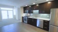 This single-floor space showcases a thoughtfully designed interior with a strong emphasis on modern aesthetics. The open-concept kitchen serves as a central highlight, featuring rich dark cabinetry that contrasts beautifully with crisp white countertops and a light-toned tile backsplash. Stainless steel appliances, including a gas range, built-in microwave, and dishwasher, lend the kitchen a polished, professional feel. A stainless steel undermount sink sits beneath a sleek faucet, completing the functional and stylish workspace. Recessed lighting illuminates the kitchen area, enhancing the clean lines and contemporary finishes. Large-format floor tiles in a neutral tone extend throughout the space, creating a seamless and cohesive look underfoot. The layout transitions smoothly from the kitchen into an adjoining open living area, which benefits from natural light streaming in through generously sized windows. The overall ambiance is clean, modern, and airy, with a neutral color palette of whites and grays that provides a versatile backdrop. The space is well-suited for those who appreciate contemporary design combined with practical, efficient living.
