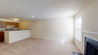 This single-floor unit offers a practical and comfortable layout designed for everyday living. The open-concept living area is generously sized, featuring neutral beige carpet, cream-toned walls, and a textured ceiling that adds subtle visual interest. Recessed lighting provides focused illumination, while a large window allows natural light to fill the space.

The kitchen is positioned adjacent to the living area and separated by a convenient breakfast bar, creating a natural division while maintaining an open feel. It is equipped with warm oak cabinetry, a coil-top electric range, a full-size refrigerator, and a range hood. Laminate wood-look flooring runs throughout the kitchen, adding a contrasting warmth to the space.

The bedroom is a well-proportioned room with soft carpeting and a wide window fitted with blinds, inviting ample daylight and outdoor views. A doorway leads directly to a closet or hallway area, enhancing the room's functionality.

The bathroom features the same wood-look laminate flooring as the kitchen, warm wood cabinetry with an integrated vanity and mirror, and a grab bar along the wall — a practical accessibility feature. The overall design of the unit is cohesive, with consistent neutral tones and functional finishes that offer a clean, move-in-ready aesthetic.