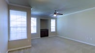 This single-floor space offers a welcoming and functional layout designed for comfortable everyday living. The main living area is generously proportioned, featuring plush wall-to-wall carpeting that adds warmth underfoot. Crown molding runs along the perimeter of the ceiling, lending a refined, classic touch to the interior. A ceiling fan with an integrated light fixture is centrally positioned, ensuring year-round comfort and efficient air circulation throughout the room. Natural light flows in abundantly through multiple windows dressed with horizontal blinds, creating a bright and airy atmosphere. The neutral, warm-toned walls complement the light carpeting, providing a versatile backdrop that suits a variety of furnishing styles. A distinctive architectural nook is built into one wall, housing a dark wood cabinet with ample storage — a practical and visually interesting design element that anchors the space. The white baseboards throughout add a crisp, clean contrast to the warm wall tones. Overall, the space presents a well-balanced combination of classic architectural detailing and practical features, making it a comfortable and inviting environment.