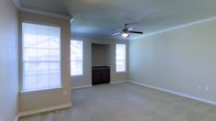 This single-floor space offers a welcoming and functional layout designed for comfortable everyday living. The main living area is generously proportioned, featuring plush wall-to-wall carpeting that adds warmth underfoot. Crown molding runs along the perimeter of the ceiling, lending a refined, classic touch to the interior. A ceiling fan with an integrated light fixture is centrally positioned, ensuring year-round comfort and efficient air circulation throughout the room. Natural light flows in abundantly through multiple windows dressed with horizontal blinds, creating a bright and airy atmosphere. The neutral, warm-toned walls complement the light carpeting, providing a versatile backdrop that suits a variety of furnishing styles. A distinctive architectural nook is built into one wall, housing a dark wood cabinet with ample storage — a practical and visually interesting design element that anchors the space. The white baseboards throughout add a crisp, clean contrast to the warm wall tones. Overall, the space presents a well-balanced combination of classic architectural detailing and practical features, making it a comfortable and inviting environment.